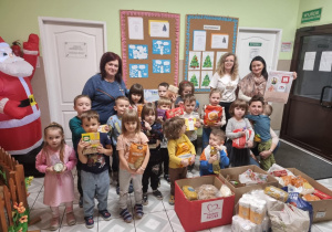 Na zdjęciu przedszkolaki wraz z Panią dyrektor i pozostałym gronem pedagogicznym stoją przy zebranej żywności. Przedszkolaki trzymają wybrane artykuły.