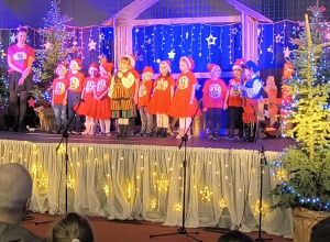 Na zdjęciu dzieci podczas występu, ubrane w czerwone bluzki z logo przedszkola i mikołajowe czapki