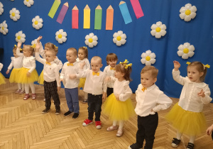 Na zdjęciu dzieci ubrane w białe bluzki, dziewczynki żółte spódniczki chłopcy ciemne spodnie i żółte muchy, podczas występu