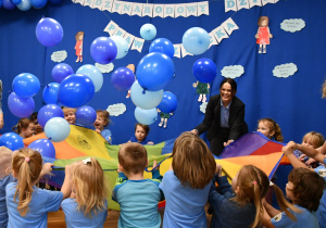 Na zdjęciu dzieci wraz z panią mecenas trzymają chustę animacyjną i huśtają niebieskie balony.