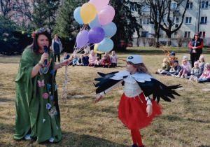Na zdjęciu Pani Wiosna trzyma w ręce balony, wokół niej biega dziewczynka w stroju bociana