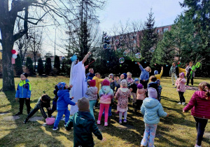 Na zdjęciu Pani Zima, puszczająca duże bańki mydlane, wokół niej dzieci łapiące bańki