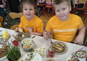 Na zdjęciu dzieci z grupy słoneczniki w podkoszulkach w kolorze pomarańczu, przy stole podczas obiadu